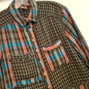 May include: A long-sleeved plaid shirt with a patchwork design in shades of black, blue, and pink. The shirt has a button-up front and a frayed hem.