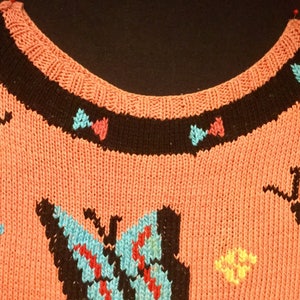 K&ouml;nnte beinhalten: Ein rosa gestrickter Pullover mit schwarzem Besatz und bunten Schmetterlingsmotiven. Die Schmetterlinge sind aus verschiedenen Farben Garn gefertigt, darunter Blau, Gelb, Rot und Lila.