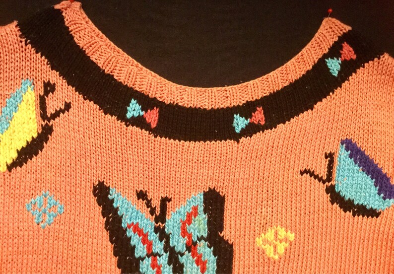 K&ouml;nnte beinhalten: Ein rosa gestrickter Pullover mit schwarzem Besatz und bunten Schmetterlingsmotiven. Die Schmetterlinge sind aus verschiedenen Farben Garn gefertigt, darunter Blau, Gelb, Rot und Lila.