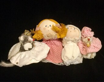 May include: Three handmade fabric dolls, one with a crown, one with a pink polka dot dress, and one with a white dress. The dolls are sitting on a black surface.