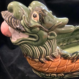 May include: A green and brown ceramic dragon planter with a wide open mouth and a pink tongue. The dragon is in a boat shape with scales on the bottom.