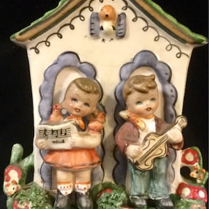May include: A ceramic wall decor featuring a white house with a blue roof and two children, a girl holding sheet music and a boy playing a violin. The house has a birdhouse on the roof with a bird inside. The children are standing on a green grassy area with red and white mushrooms.