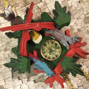 May include: A wooden cuckoo clock with a red and green design. The clock face has Roman numerals and a blue bird with a yellow beak. The clock is hanging from a gold chain.
