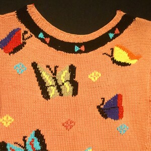 K&ouml;nnte beinhalten: Ein rosa gestrickter Pullover mit einem bunten Schmetterlingsmuster. Der Pullover hat kurze &Auml;rmel und einen runden Ausschnitt. Die Schmetterlinge sind in verschiedenen Farben gestickt, darunter Rot, Blau, Gelb, Gr&uuml;n und Schwarz.