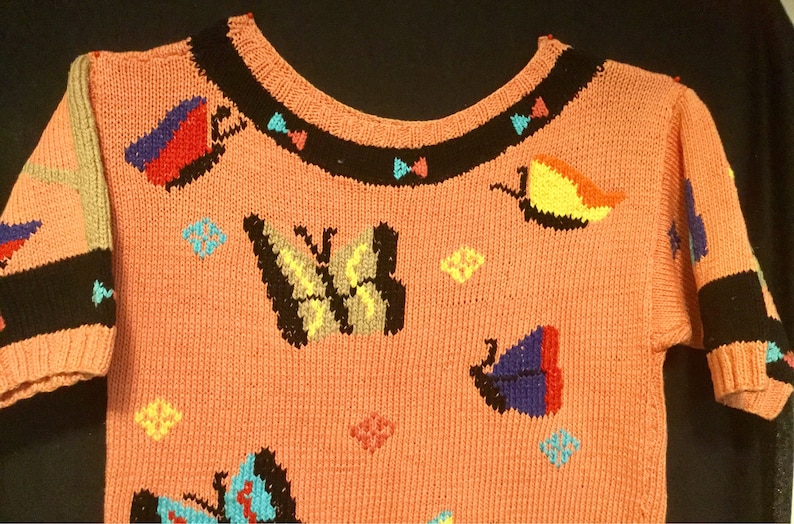 K&ouml;nnte beinhalten: Ein rosa gestrickter Pullover mit einem bunten Schmetterlingsmuster. Der Pullover hat kurze &Auml;rmel und einen runden Ausschnitt. Die Schmetterlinge sind in verschiedenen Farben gestickt, darunter Rot, Blau, Gelb, Gr&uuml;n und Schwarz.