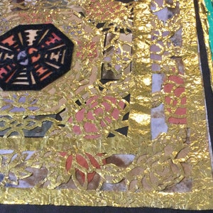 May include: A close-up of a fabric with a gold leaf design. The fabric has a geometric pattern with a black center and red and orange accents. The gold leaf is applied in a layered pattern, creating a textured effect.