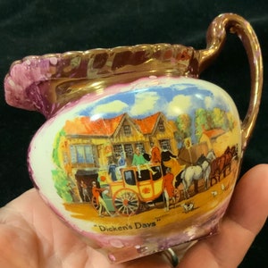 May include: A small, white ceramic creamer with a pink and gold glaze. It features a painted scene of a horse-drawn carriage and a building with the text "Dickens' Days".