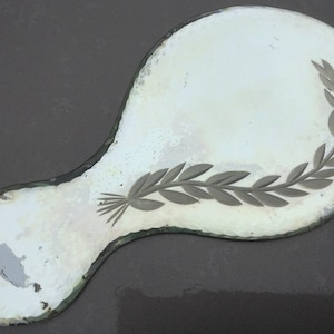 Pu&ograve; includere: Uno specchio da mano antico di forma ovale con una superficie in vetro argentato. Lo specchio presenta un motivo decorativo inciso di un ramo frondoso sul lato destro.