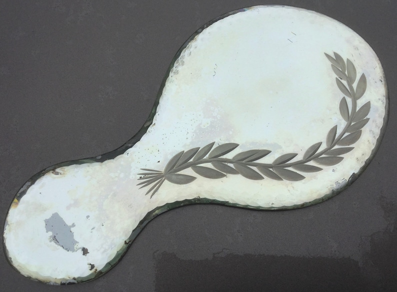 Pu&ograve; includere: Uno specchio da mano antico di forma ovale con una superficie in vetro argentato. Lo specchio presenta un motivo decorativo inciso di un ramo frondoso sul lato destro.