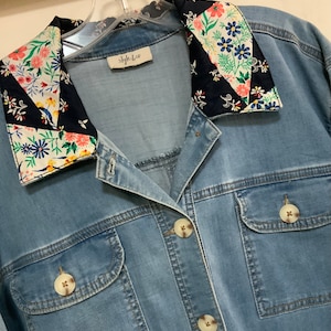 May include: A light blue denim jacket with a floral patterned collar. The collar is made of a black and white fabric with a floral print. The jacket has two pockets on the front and a button closure.