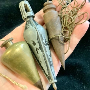 May include: Three vintage plumb bobs, one brass, one silver, and one wood, with string attached to the wood plumb bob.