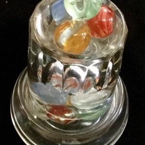 May include: A glass jar with a clear base and a textured glass top. The jar contains a collection of colorful marbles, including green, blue, red, and orange.