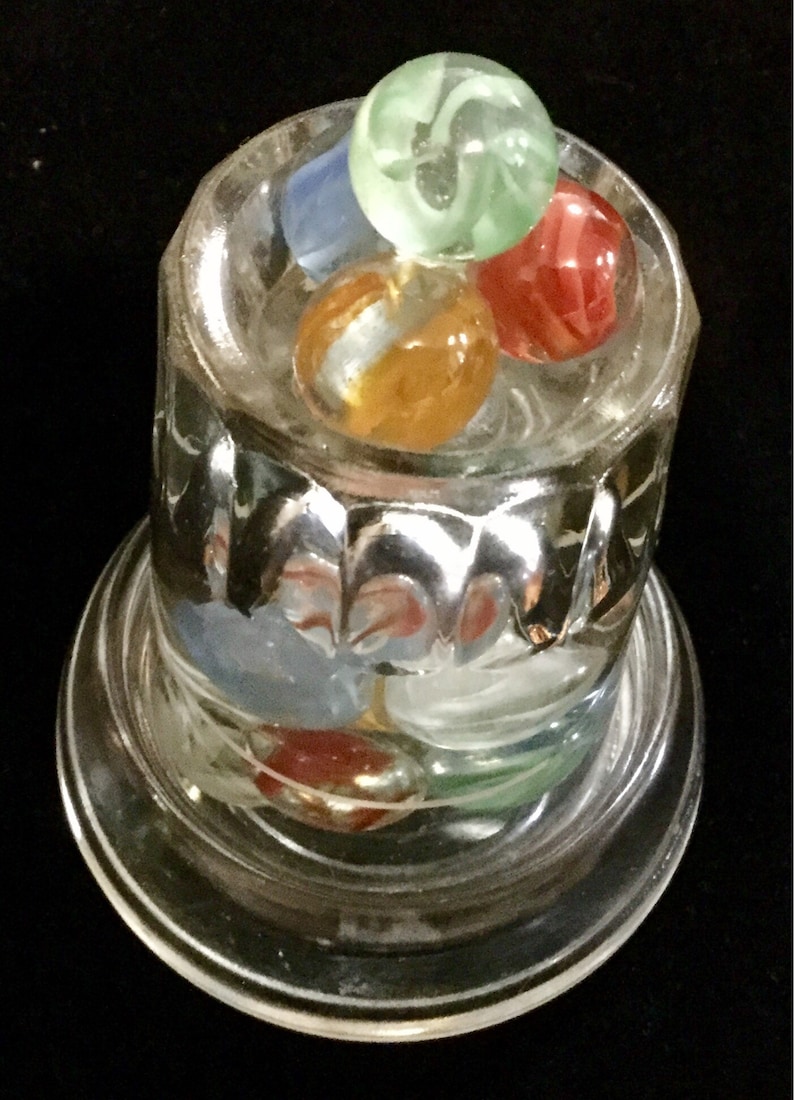 May include: A glass jar with a clear base and a textured glass top. The jar contains a collection of colorful marbles, including green, blue, red, and orange.