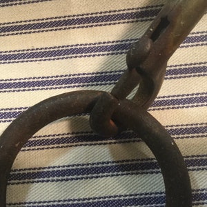 May include: A rusty metal hook with a ring attached to the end. The hook is bent at a 90-degree angle and has a rough, textured surface.