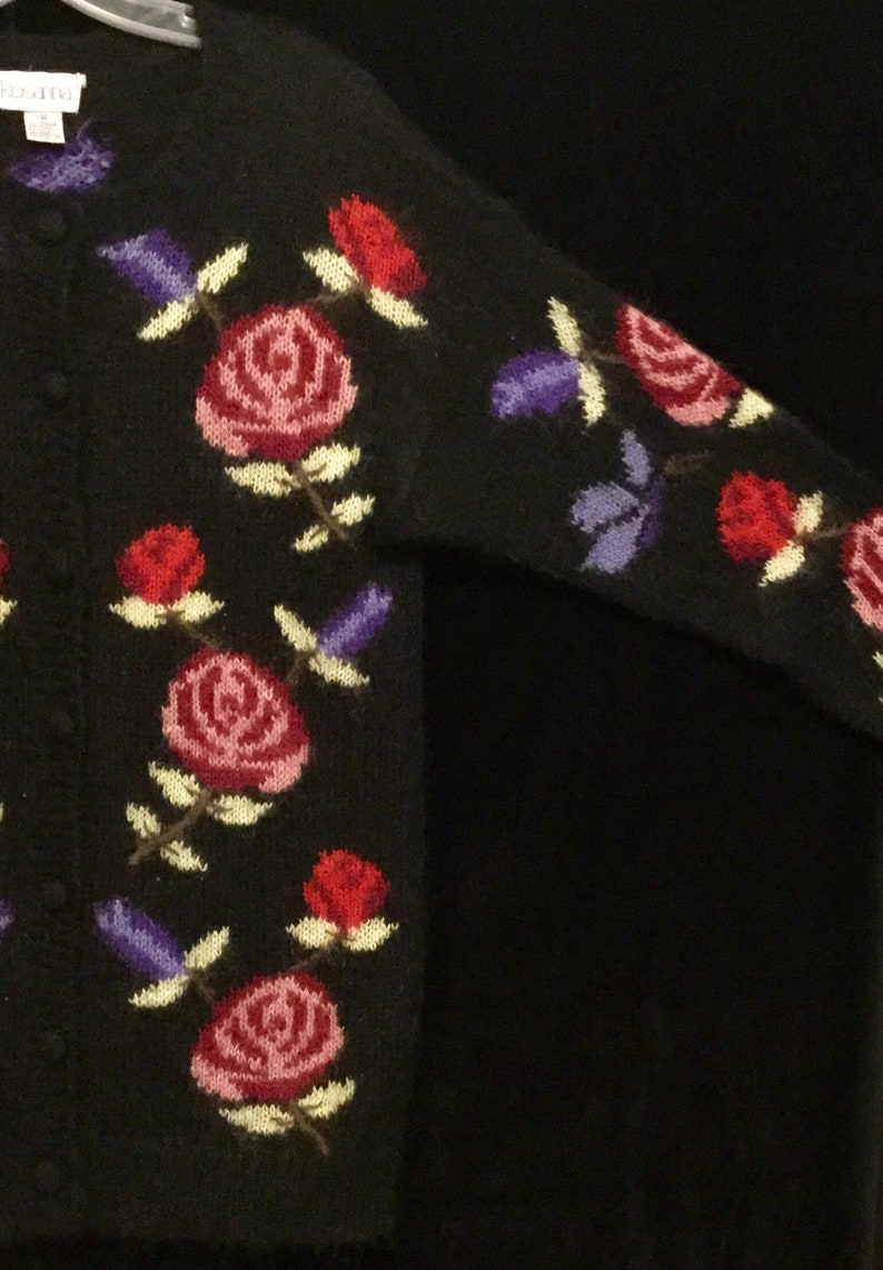 May include: Black cardigan sweater with a floral pattern of pink, red, and purple roses. The sweater has a button closure.