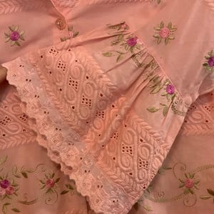 May include: A pale pink blouse with detailed floral embroidery and cutwork. The sleeves have a ruffled cuff with delicate openwork patterns. Small pink and green floral embroidery accents the fabric, creating a delicate design.