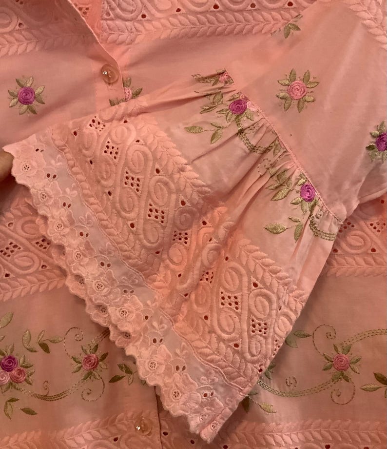 May include: A pale pink blouse with detailed floral embroidery and cutwork. The sleeves have a ruffled cuff with delicate openwork patterns. Small pink and green floral embroidery accents the fabric, creating a delicate design.