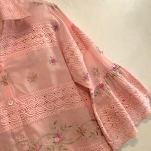 May include: A pale pink shirt with detailed embroidery and eyelet patterns. The shirt has floral embroidery in pink and green, with decorative patterns along the sleeves and body.