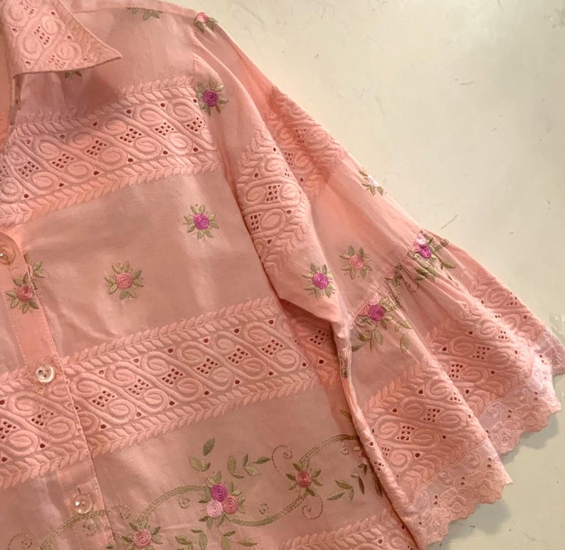 May include: A pale pink shirt with detailed embroidery and eyelet patterns. The shirt has floral embroidery in pink and green, with decorative patterns along the sleeves and body.