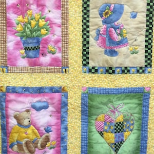 May include: A yellow and pink quilt with four squares. Each square has a different design. The top left square has a bouquet of yellow flowers in a blue pot. The top right square has a girl in a blue dress and a pink hat. The bottom left square has a brown teddy bear wearing a yellow shirt. The bottom right square has a patchwork heart with a green background.