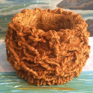 May include: A brown, woven, coconut coir basket with a round shape and a textured surface. The basket is made of natural fibers and has a rustic look.