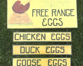 Puede incluir: Un letrero de madera amarillo con el texto "FREE RANGE EGGS" en letras negras. Debajo del letrero hay tres letreros más pequeños, cada uno con un tipo de huevo diferente: "CHICKEN EGGS", "DUCK EGGS" y "GOOSE EGGS".