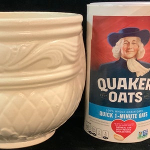 May include: A white ceramic planter with a decorative design. The planter has a wide base and a narrow top. The planter is next to a box of Quaker Oats Quick 1-Minute Oats. The box features the Quaker Oats mascot, a man in a blue coat and white wig, with the text "Quaker Oats" and "100% Whole Grain Oats".