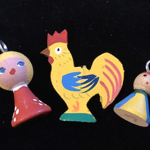May include: Three wooden ornaments, a rooster, a girl, and a boy, painted in bright colors. The rooster is yellow with blue and red accents, the girl is pink with red and white accents, and the boy is brown with yellow and blue accents.