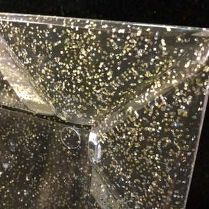 May include: A clear plastic tray with a gold and silver glitter finish. The tray has a square shape with angled edges.