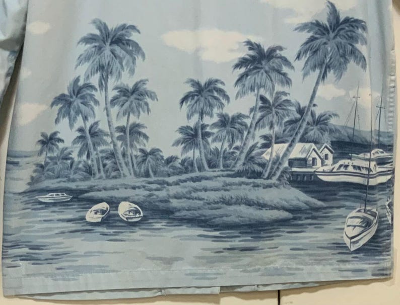 May include: Light blue button-down shirt featuring a tropical island scene. The print includes palm trees, boats, and small buildings. The shirt has a classic collar and long sleeves.