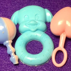 Pode incluir: Tr&ecirc;s brinquedos para beb&eacute;s: um chocalho azul e rosa, um anel azul com a cara de um c&atilde;o de desenho animado e um mordedor rosa em forma de cora&ccedil;&atilde;o.