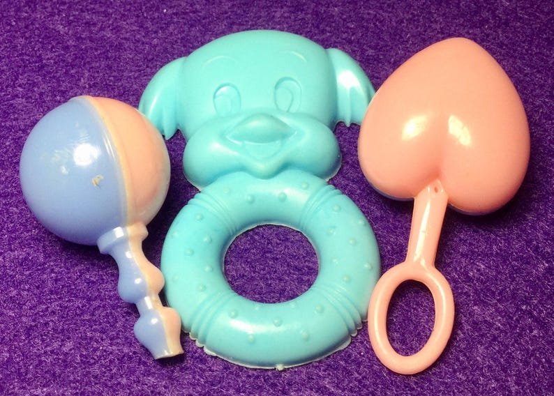 Pode incluir: Tr&ecirc;s brinquedos para beb&eacute;s: um chocalho azul e rosa, um anel azul com a cara de um c&atilde;o de desenho animado e um mordedor rosa em forma de cora&ccedil;&atilde;o.