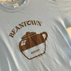 May include: A light blue t-shirt with the text "BEANTOWN" in brown letters above a brown illustration of a teapot with the word "BOSTON" printed on the side.