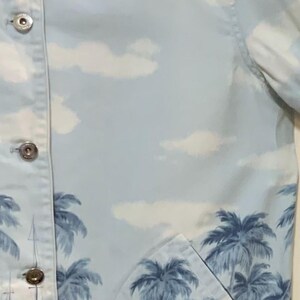 May include: Light blue button-up jacket with a tropical palm tree and ocean scene print. The jacket features a light beige collar and silver buttons. The design includes white clouds in the sky.