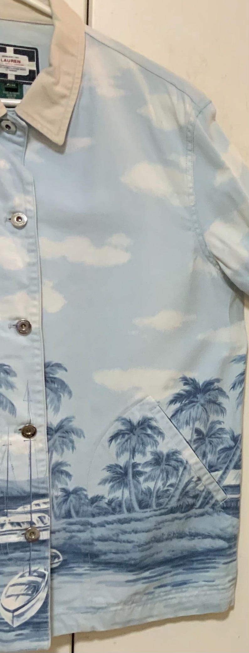 May include: Light blue button-up jacket with a tropical palm tree and ocean scene print. The jacket features a light beige collar and silver buttons. The design includes white clouds in the sky.