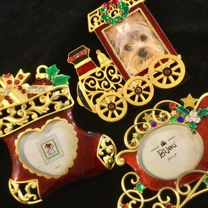 May include: Three gold-tone Christmas ornaments with red and green accents. The ornaments are shaped like a stocking, a train, and a sleigh. Each ornament has a small picture frame.