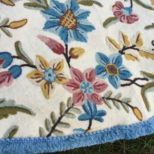 May include: A round, white tablecloth with a blue fringe border features a floral embroidery design. The embroidery includes pink, blue, and yellow flowers with green leaves.