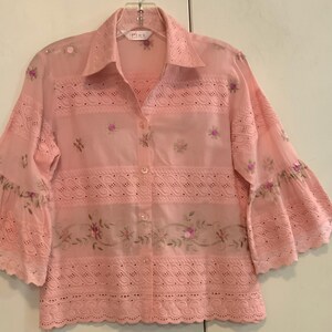 May include: A pale pink button-down shirt with eyelet and floral embroidery. The shirt features bell sleeves and a collared neckline. The embroidery includes small pink flowers and green leaves. The shirt is hanging on a clear hanger.