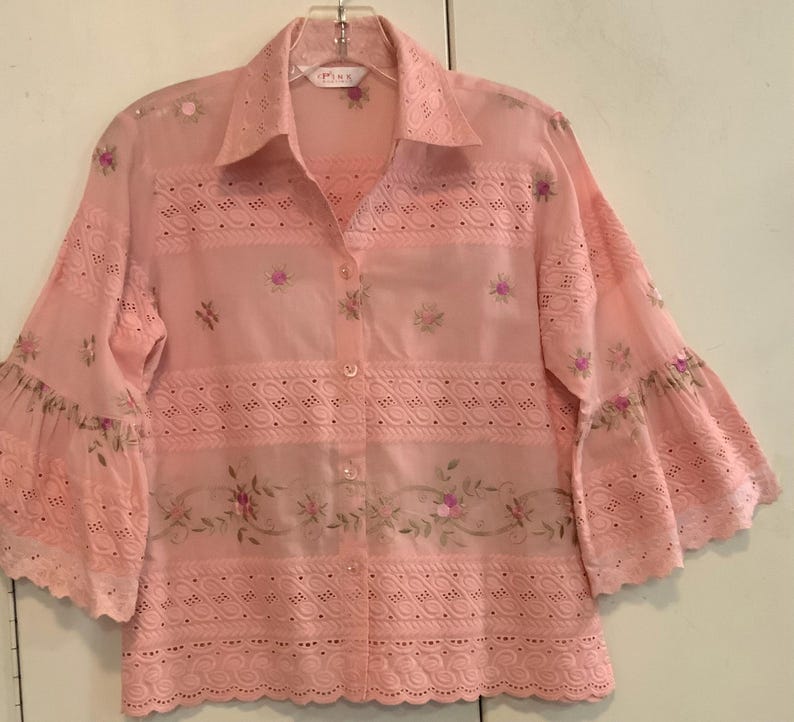 May include: A pale pink button-down shirt with eyelet and floral embroidery. The shirt features bell sleeves and a collared neckline. The embroidery includes small pink flowers and green leaves. The shirt is hanging on a clear hanger.