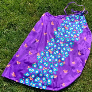 Op de afbeelding: Paarse wikkelrok met een bloemenpatroon met witte bloemen met blauwe centra en oranje accenten. De rok heeft een vlinderpatroon.