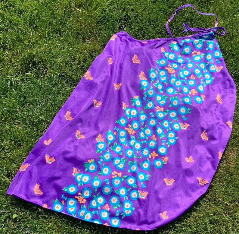 Op de afbeelding: Paarse wikkelrok met een bloemenpatroon met witte bloemen met blauwe centra en oranje accenten. De rok heeft een vlinderpatroon.
