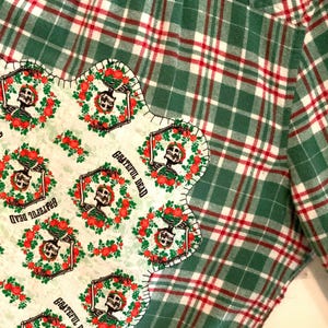 May include: A green and red plaid flannel shirt with a patch sewn on the front. The patch is a white fabric with a repeating pattern of a skeleton holding a rose wreath and the words "Grateful Dead".