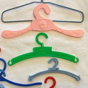 May include: A collection of plastic doll hangers in various colors, including pink, green, blue, and red. Some hangers have a decorative design, while others have text that reads "Tailored" and "Showroom Dolls".