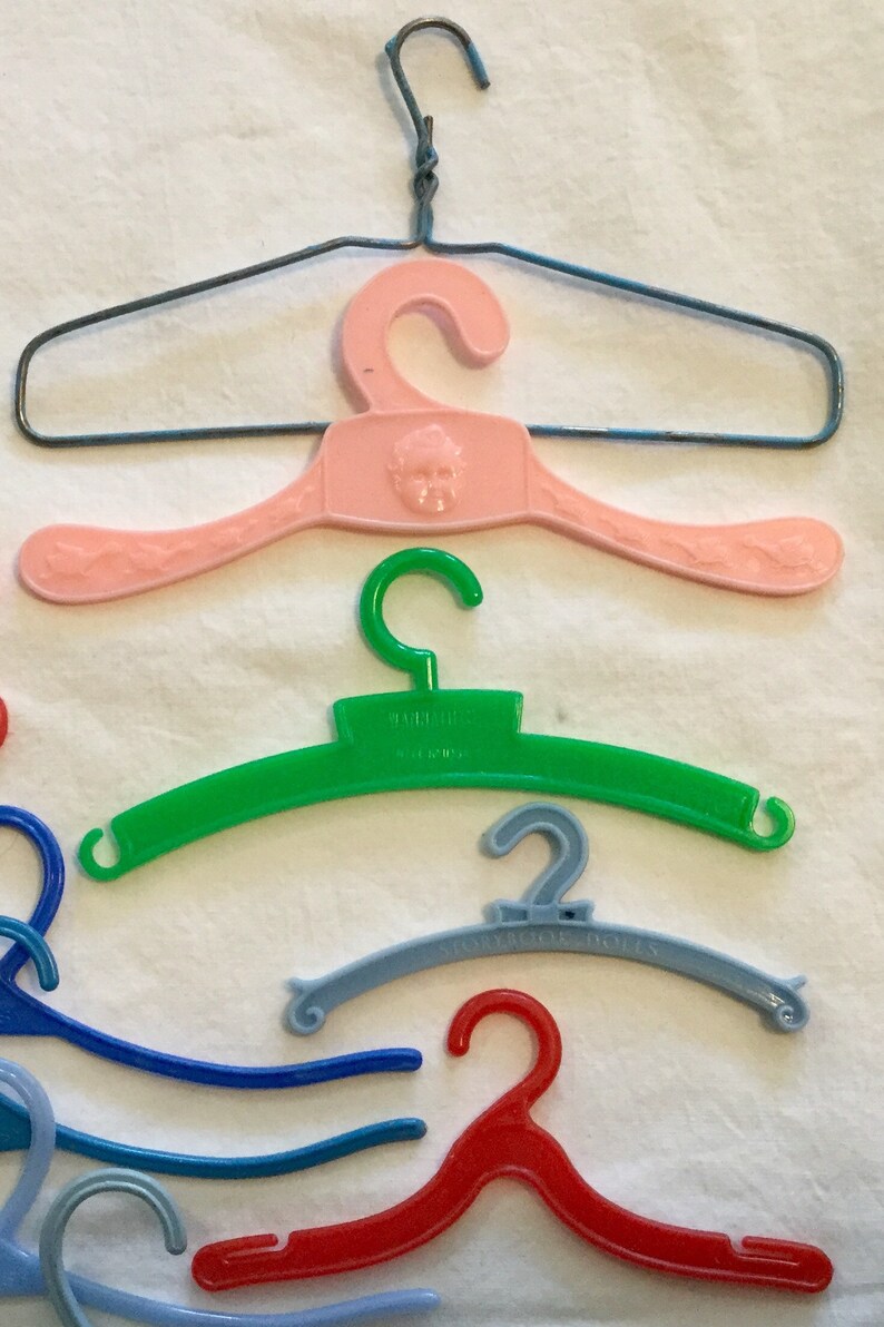 May include: A collection of plastic doll hangers in various colors, including pink, green, blue, and red. Some hangers have a decorative design, while others have text that reads "Tailored" and "Showroom Dolls".
