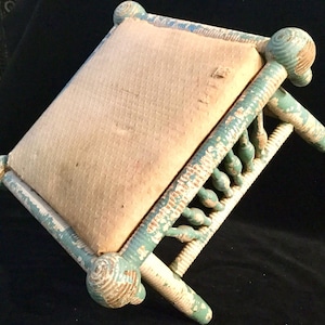 May include: An antique footstool with a distressed, painted finish. The frame is painted in a faded teal and white, with a beige, textured cushion. The legs have decorative turned spindles. A vintage piece of furniture.