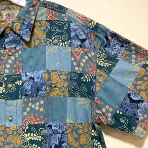 May include: A short-sleeve button-down shirt featuring a patchwork design with various blue, teal, and yellow floral and paisley patterns. The garment includes a standard collar, a front button placket with light brown buttons, and a chest pocket.