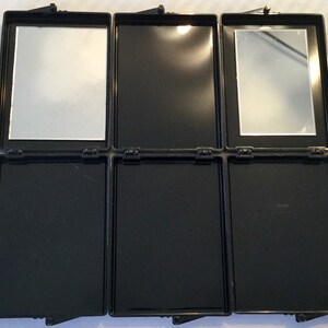 May include: A black, rectangular, three-panel compact mirror with two mirrors and three empty compartments. The compact is open, revealing the mirrors and compartments. The mirrors are rectangular and set in the top panels. The compact has a hinged closure.