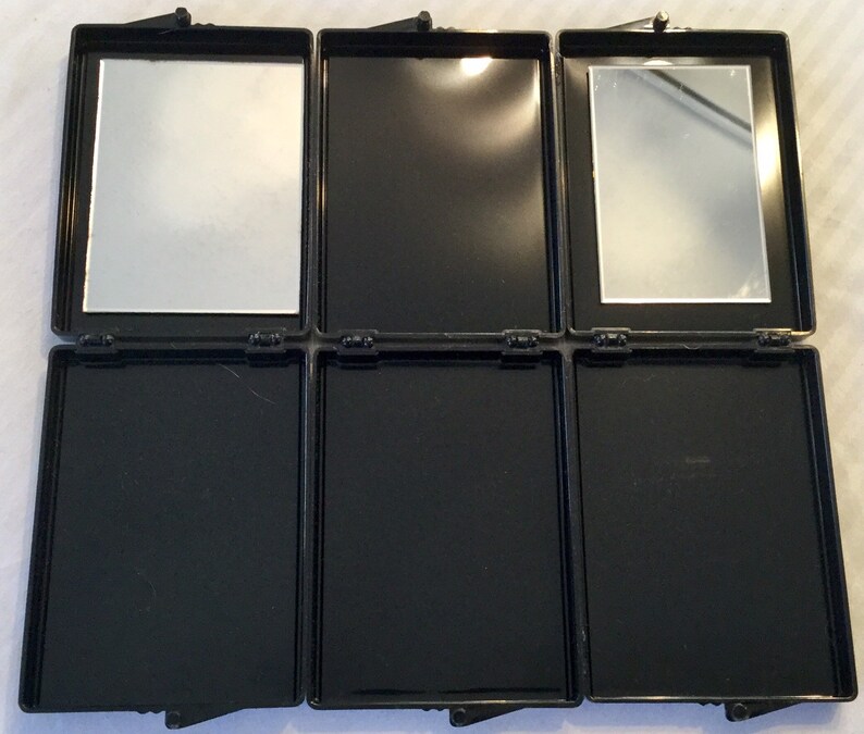 May include: A black, rectangular, three-panel compact mirror with two mirrors and three empty compartments. The compact is open, revealing the mirrors and compartments. The mirrors are rectangular and set in the top panels. The compact has a hinged closure.