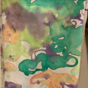 May include: Close-up of fabric with an abstract watercolor design. The fabric features a white base with vibrant splashes of green, purple, and orange hues. The edges of the fabric are visible, with a stitched seam.