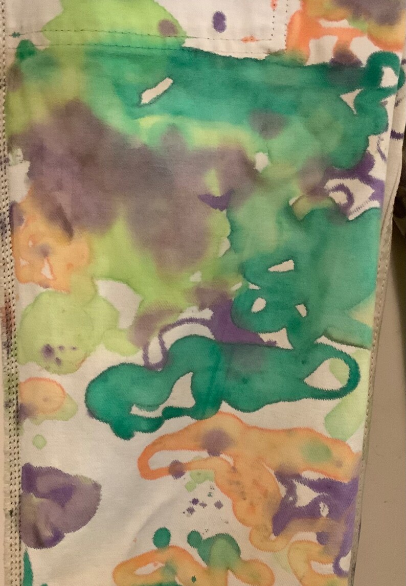 May include: Close-up of fabric with an abstract watercolor design. The fabric features a white base with vibrant splashes of green, purple, and orange hues. The edges of the fabric are visible, with a stitched seam.